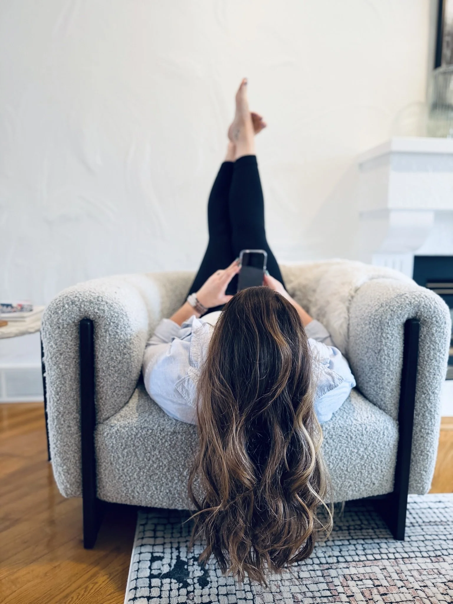 Woman with long wavy hair lying on a couch, holding a phone, with her legs raised and resting against a white wall.