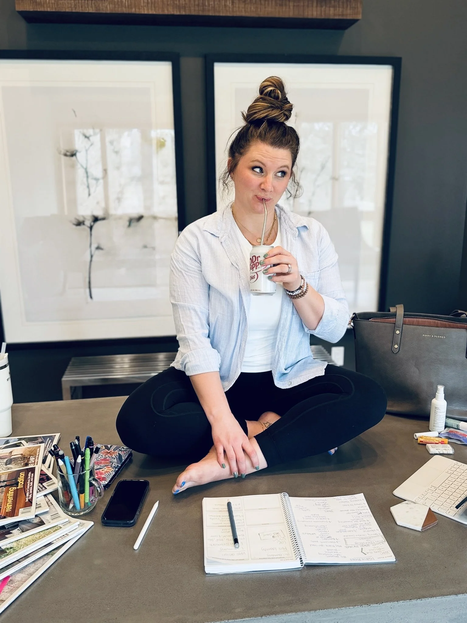 A woman with a top knot hairstyle sitting cross-legged on a table, sipping from a Dr Pepper can with a straw, surrounded by office supplies and a bag.