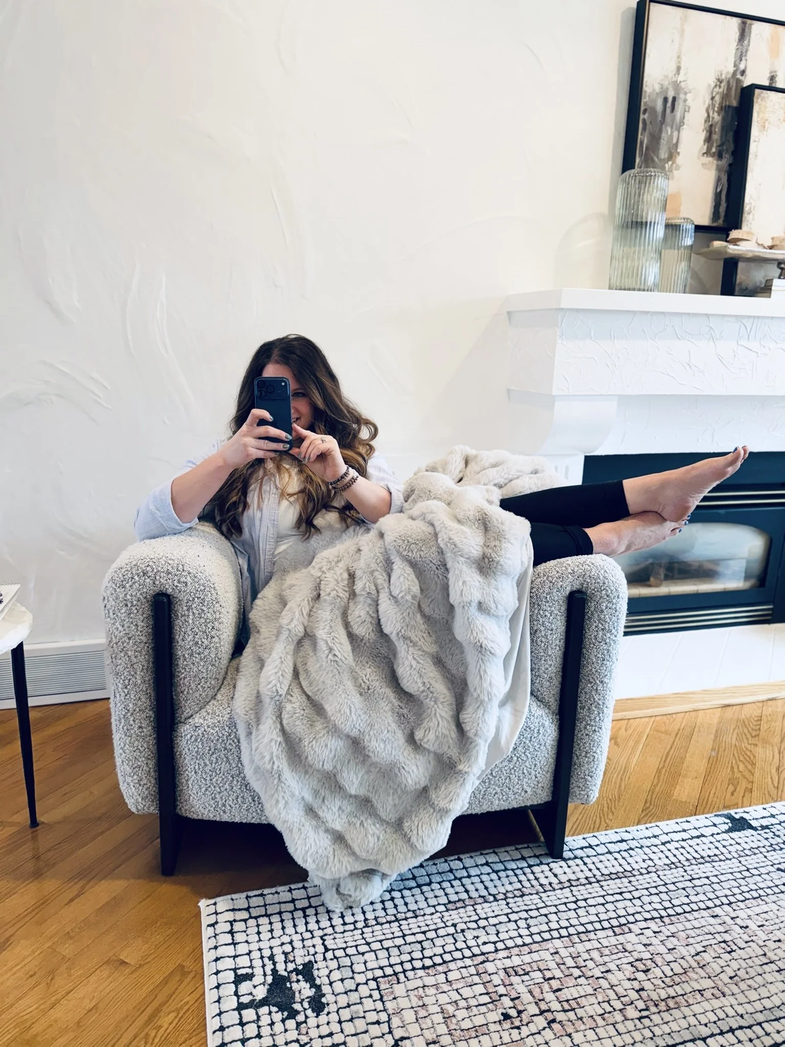 Woman sitting in a cozy living room taking a selfie with her phone, draped in a fluffy blanket, with her legs extended on the armrest of the chair.