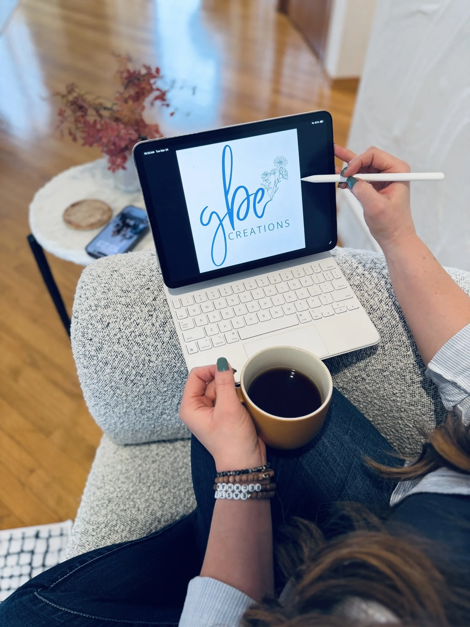 Person sitting on a couch with a tablet displaying 'Jbe Creations' logo, holding a cup of coffee, with a small table nearby holding a phone, a cookie, and a small bouquet of pink flowers.