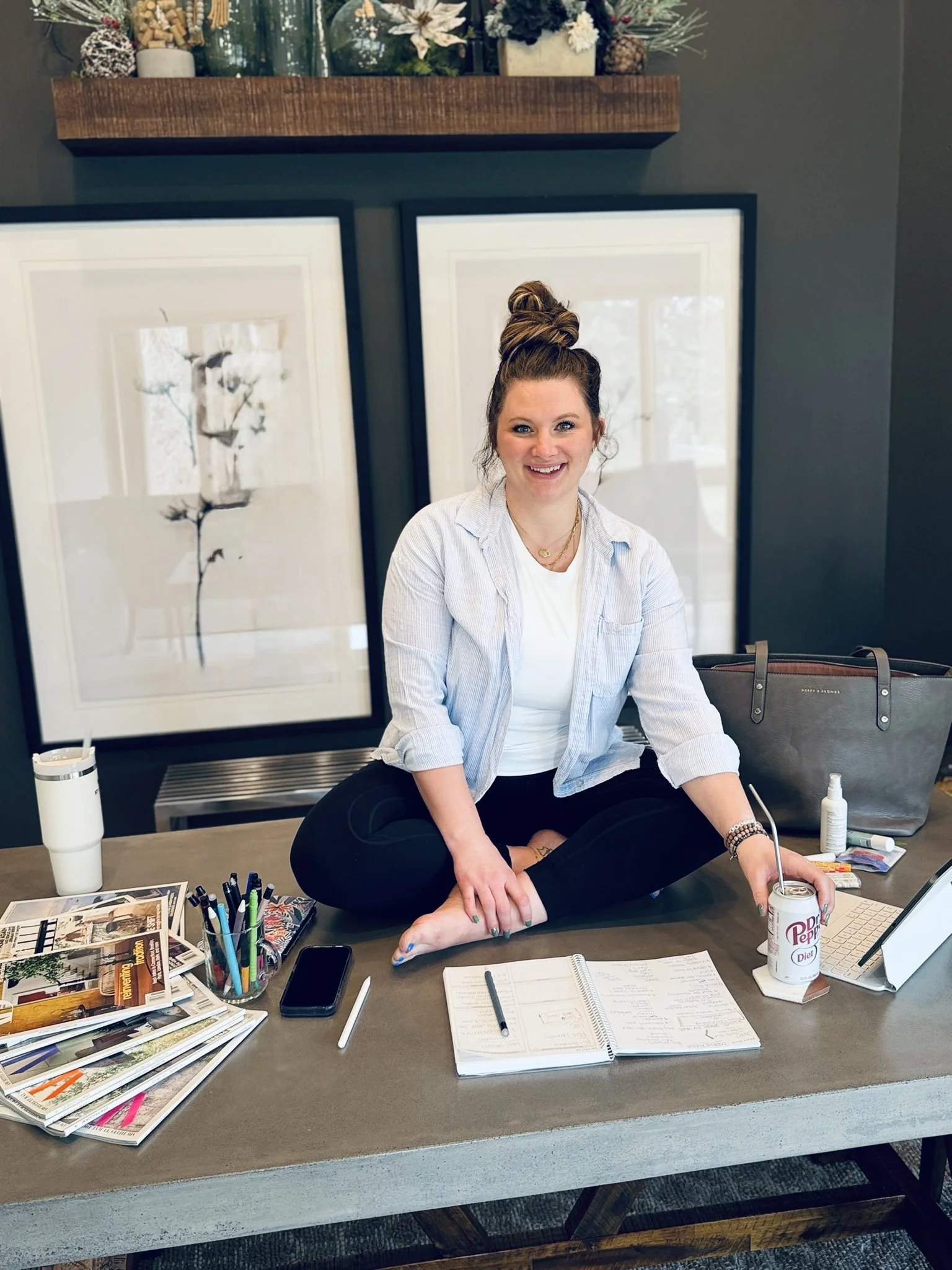 A smiling woman with her hair in a bun, sitting on a table in a cozy indoor setting, surrounded by art supplies, notebooks, a laptop, a soda can with a straw, and personal items.