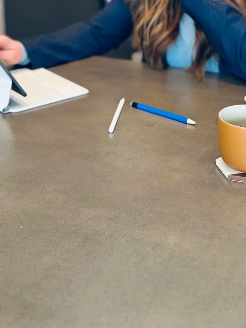 A table with a cup, a white and blue pen, a white pen, a beige notebook, and a person sitting with long hair and a blue jacket using a laptop.