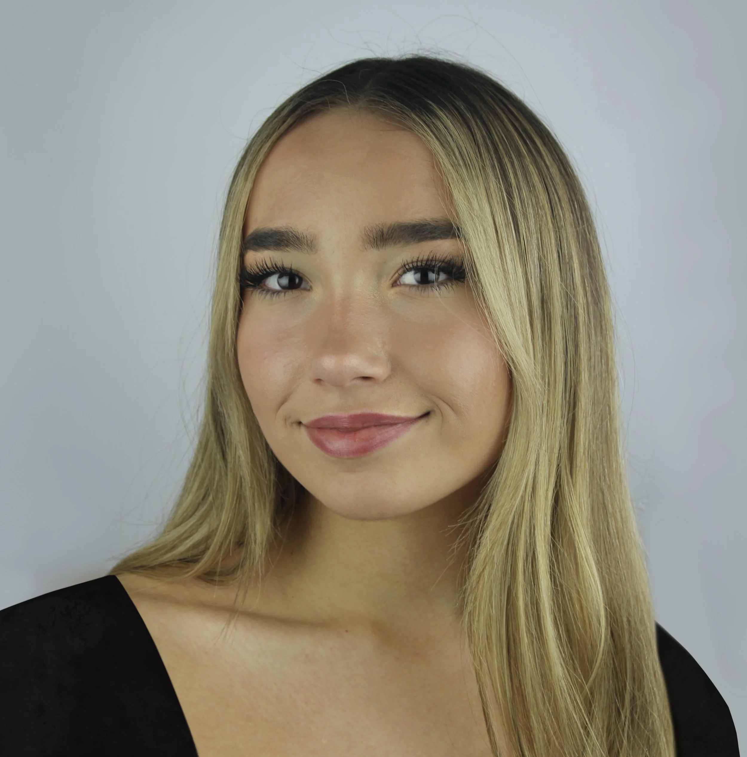 A young woman with long blonde hair, light makeup, and a gentle smile, wearing a black top, looking at the camera against a plain gray background.