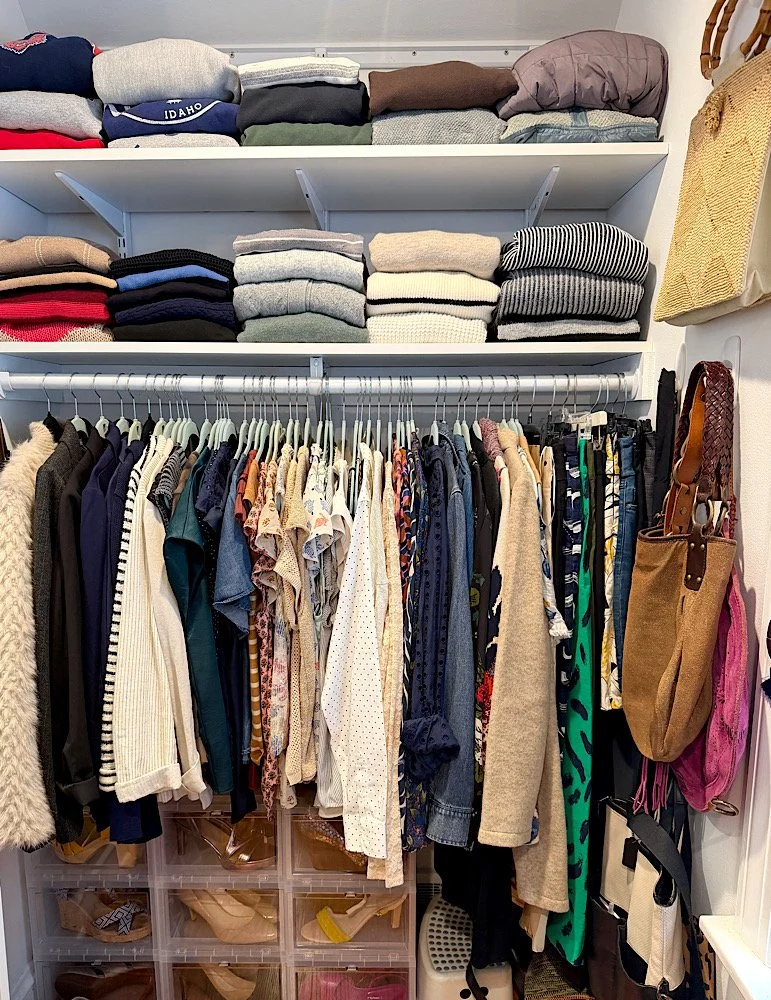 Closet with neatly folded sweaters on top shelves and hanging shirts, blouses, and jackets inside. Shoe storage at the bottom and bags hanging on the side.