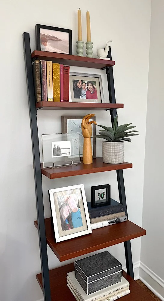 A black and wooden leaning bookshelf containing framed photos, books, candles, decorative items, a potted plant, and a storage box.
