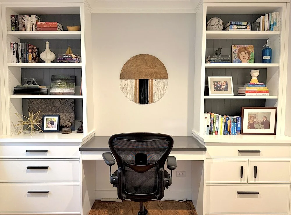 A modern home office with white built-in bookshelves on both sides, a gray desk, and an ergonomic office chair. Decor includes framed photos, books, sculptures, and a round wall art piece made of wood and textile on a white wall.