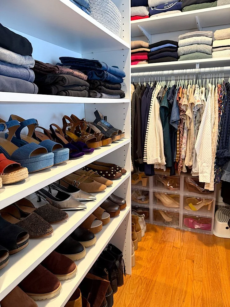 Closet with shelves holding folded clothes, shoes, and storage bins, and a hanging section with various shirts and jackets.