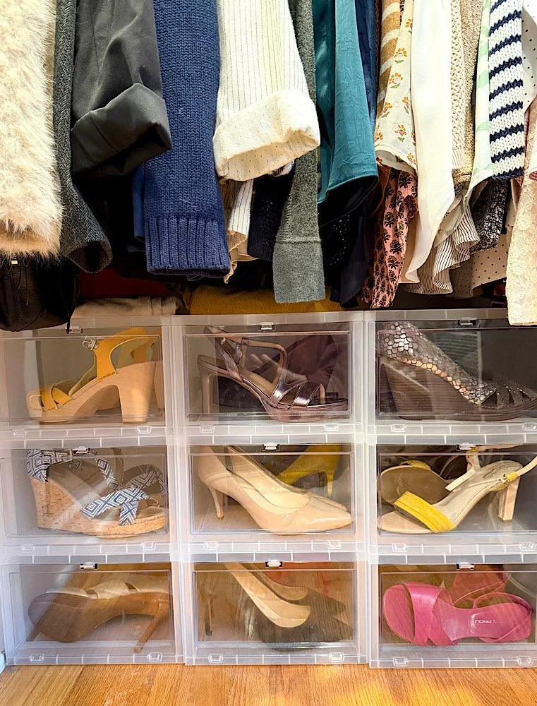 Display of women's high-heeled shoes inside clear storage containers below hanging clothing, including sweaters and blouses.