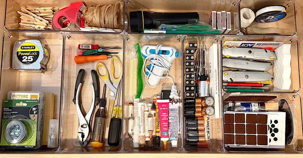 Organized storage drawer with tools, batteries, measuring tape, scissors, screwdrivers, small lighter, glue, rolls of tape, scissors, and various small hardware items.