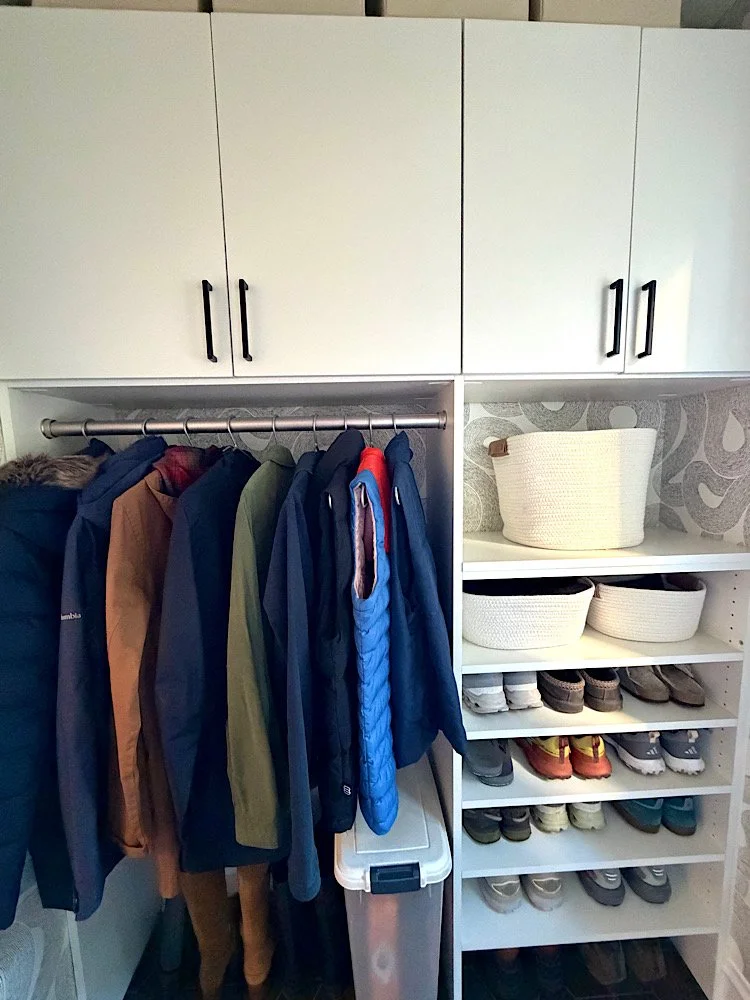 Closet with jackets on a rod, white shelves with baskets and shoes, upper cabinet with four doors.