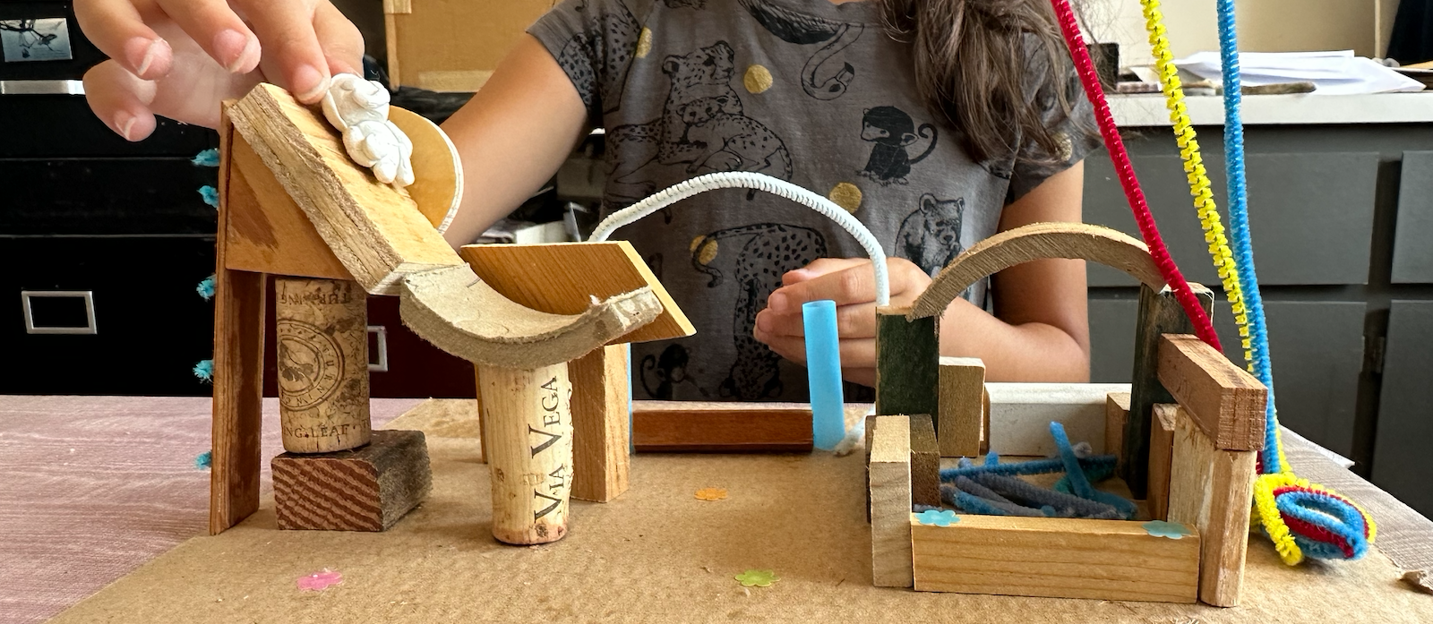 Child building a miniature wooden roller coaster with various small wooden pieces, pipe cleaners, and fabric in a classroom or workshop setting.