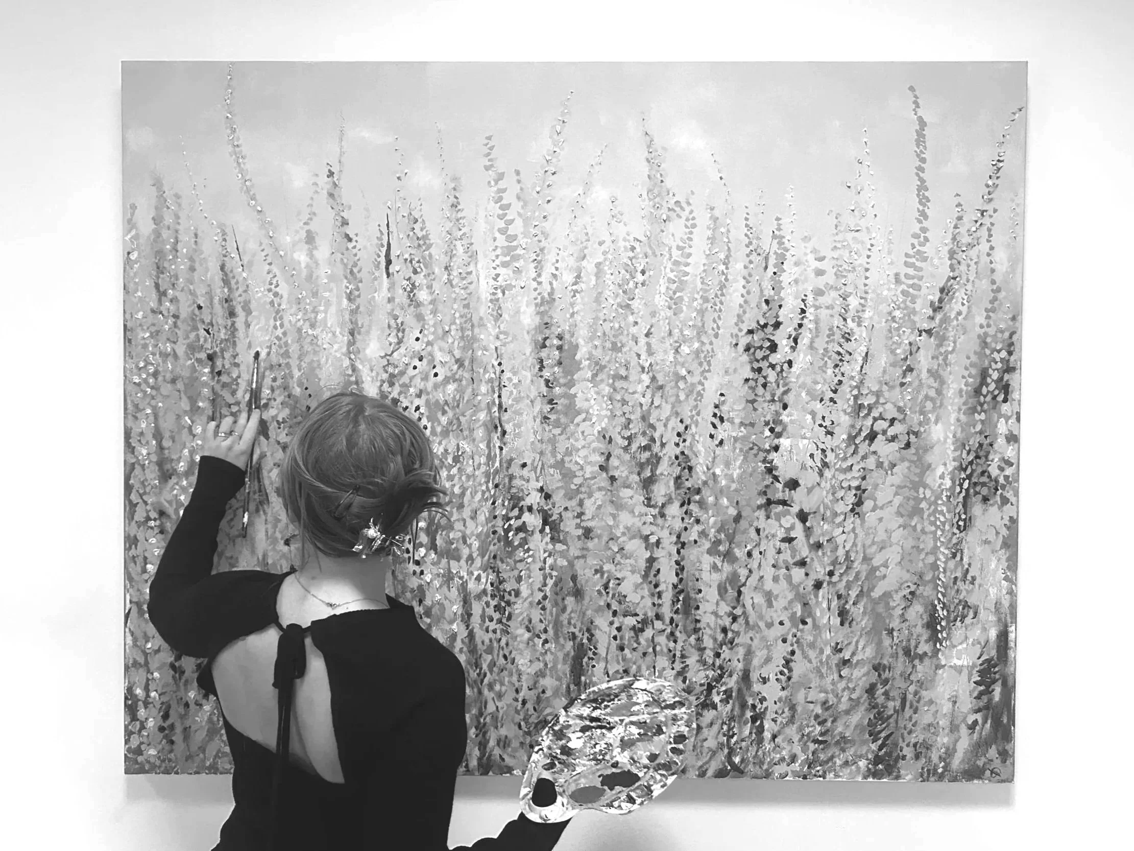 A woman with short hair, wearing a black dress with a back cutout, is standing in front of a large, abstract, textured painting. She is holding a palette and using her right hand to make a gesture near the painting.