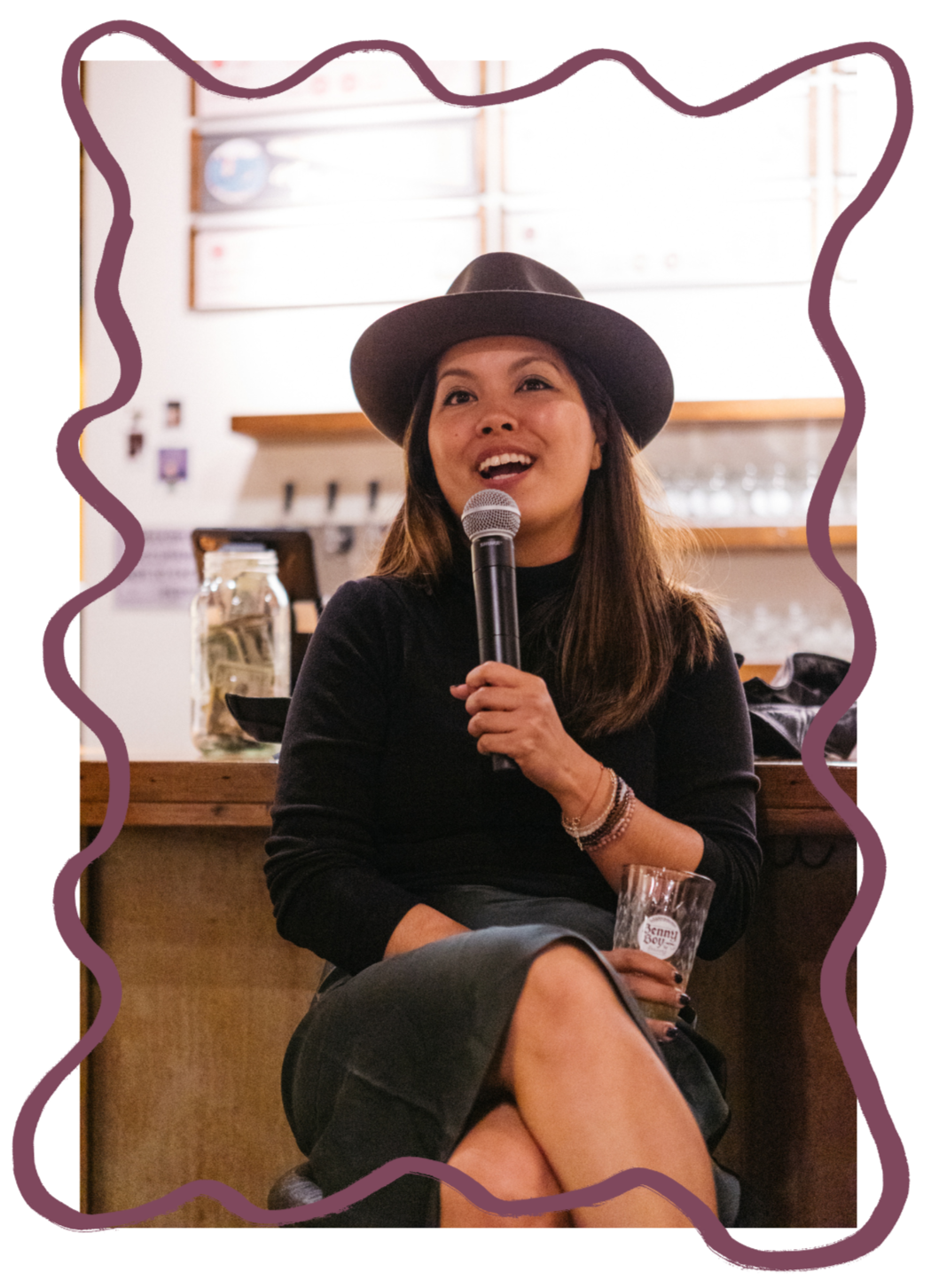 A woman with long hair wearing a hat and black shirt, sitting on a barstool, holding a microphone and a glass, smiling and possibly speaking or singing, with a blurred bar or café background.