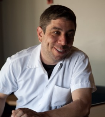 A man smiling, wearing a white short-sleeve shirt, sitting indoors.