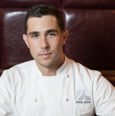Portrait of a male chef wearing a white chef uniform, sitting in front of a red padded background.