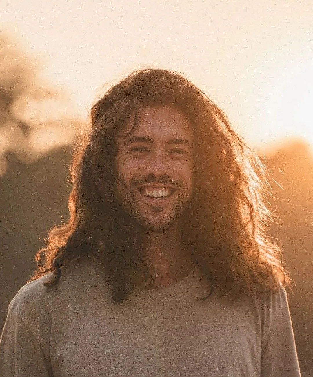 Jeune homme souriant avec de longs cheveux bruns, portant un t-shirt beige, le soleil de coucher en arrière-plan. Professeur de yoga and breathwork.