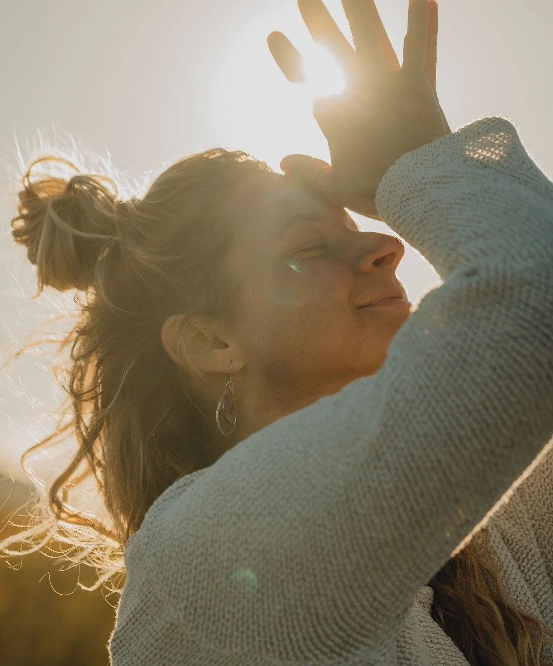 Femme souriante avec cheveux attachés en chignon, portant boucles d'oreilles, en plein soleil avec lumière dorée, levant la main devant le soleil. Méditation et yoga.