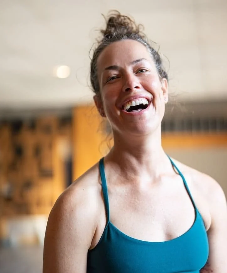 Une femme souriante, portant un maillot de sport bleu, avec des cheveux attachés en chignon, dans un environnement intérieur. Professeur de yoga.