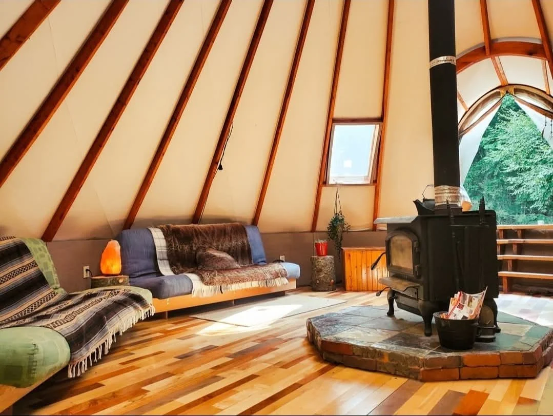 Intérieur d'une cabane en bois avec un poêle à bois sur un socle en briques, deux canapés, une petite table, et une fenêtre avec vue sur la nature.