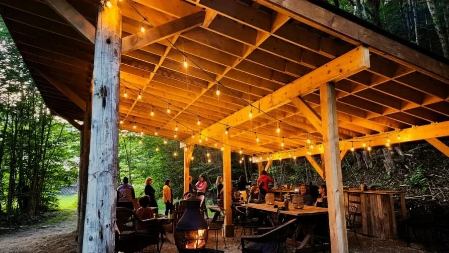 Groupe de personnes sous une grande pergola en bois, éclairée par des guirlandes lumineuses, dans un environnement forestier, lors d'une soirée.
