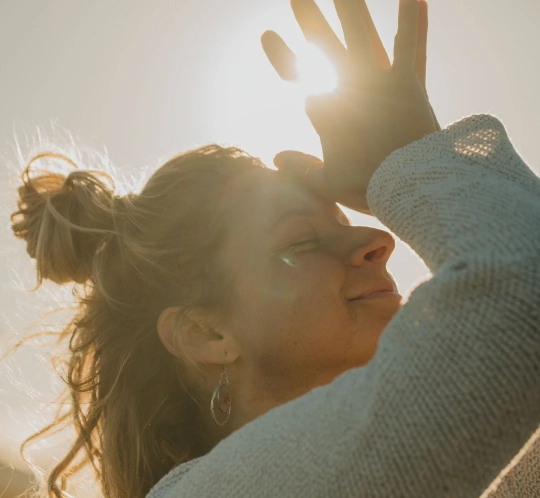 Une femme souriante avec des cheveux attachés en chignon, portant un vêtement clair, se protège du soleil avec sa main levée devant le visage, en arrière-plan un ciel clair avec la lumière du soleil.