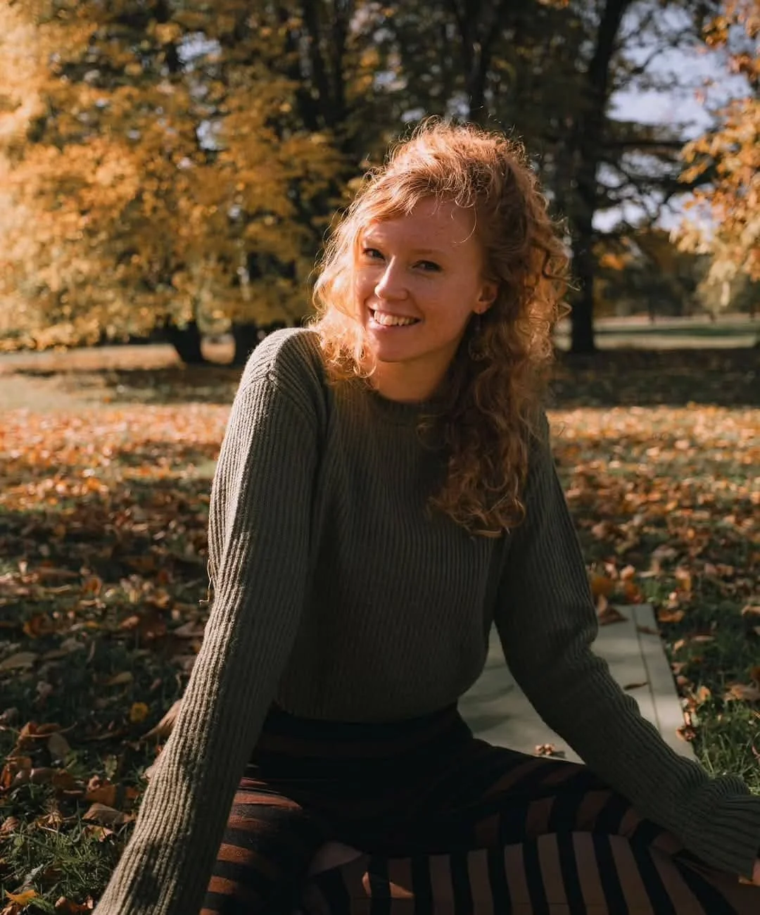 Jeune femme aux cheveux roux bouclés, souriante, assise dans un parc en automne, portant un pull gris et un pantalon à rayures. Professeur de yoga.
