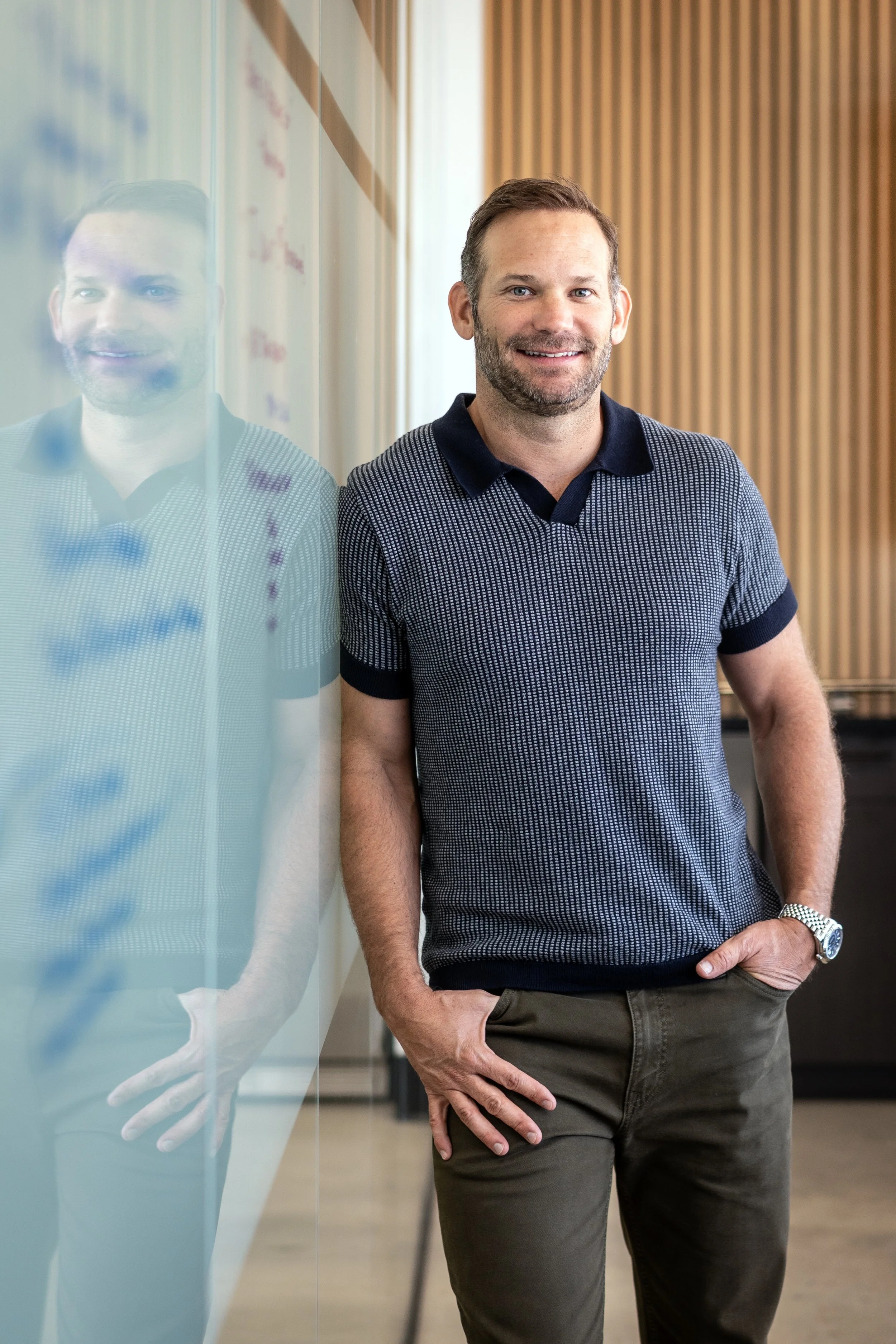 A man with a beard and short brown hair standing next to a glass wall, wearing a navy blue polo shirt with a small checkered pattern and dark pants, smiling at the camera.