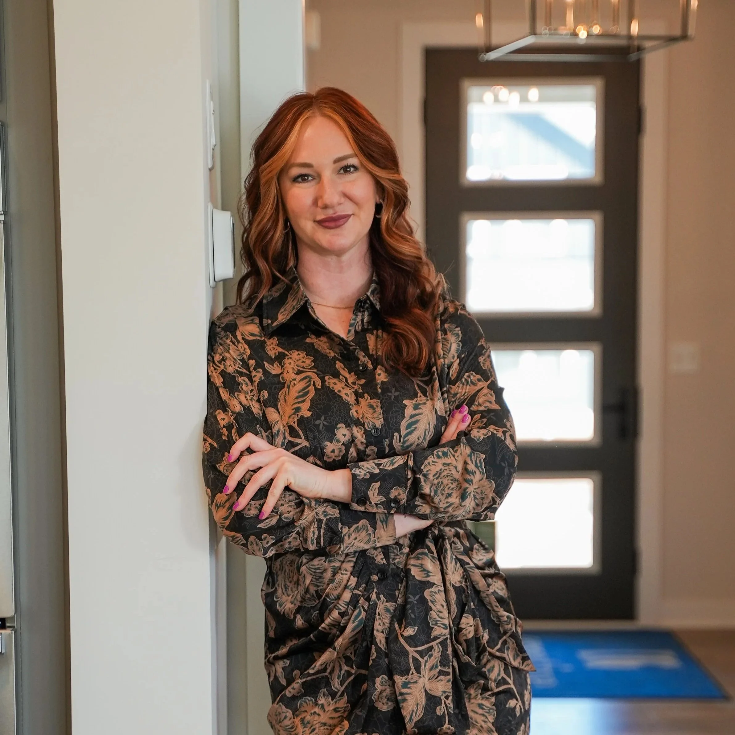 A woman with long, wavy red hair standing indoors, leaning against a wall with arms crossed, smiling at the camera, in front of a modern gray door with square glass panels.