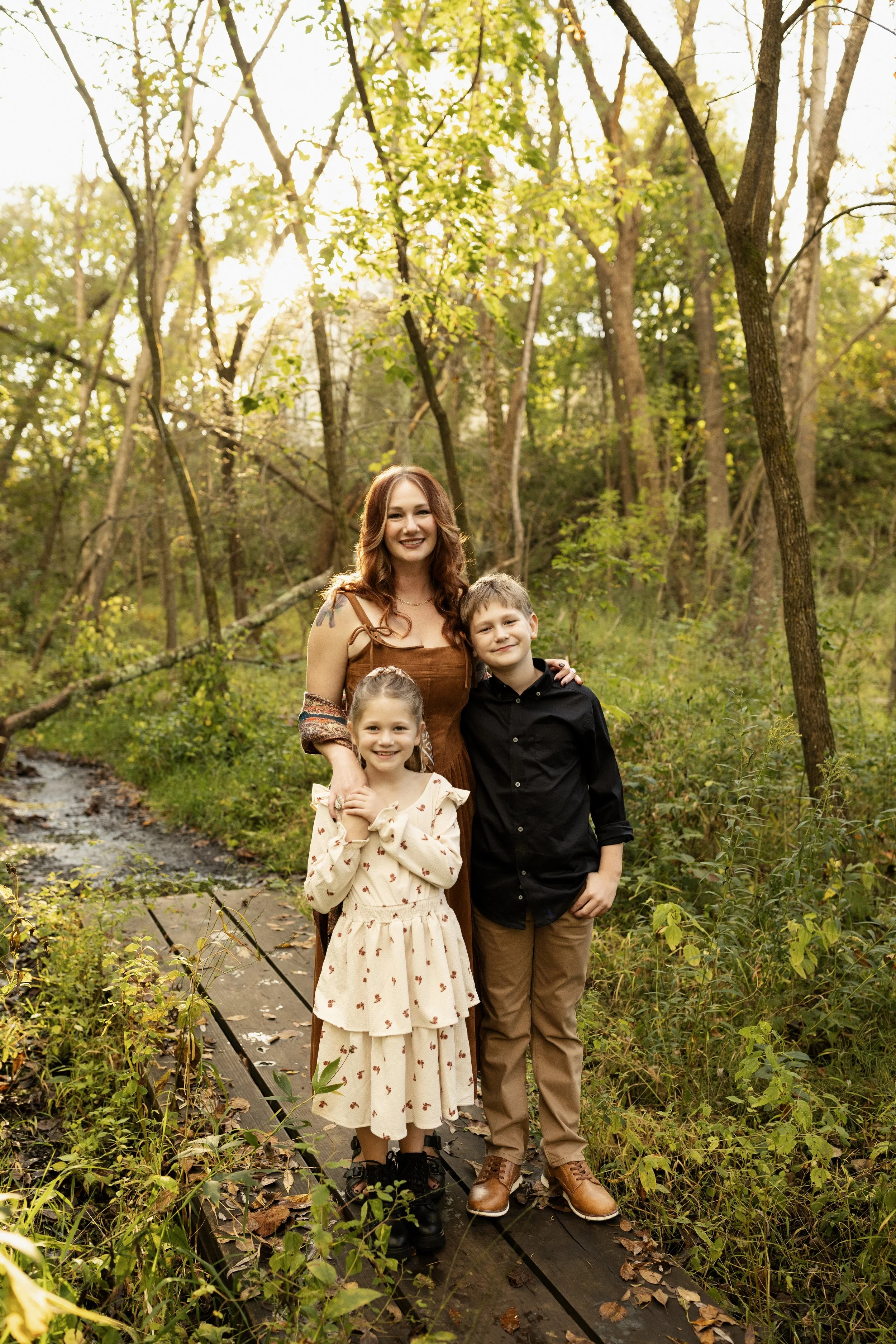 A woman with two kids standing on a forest pathway in autumn. The woman has red hair, the girl has light brown hair, and the boy has short brown hair. They are smiling and dressed casually.