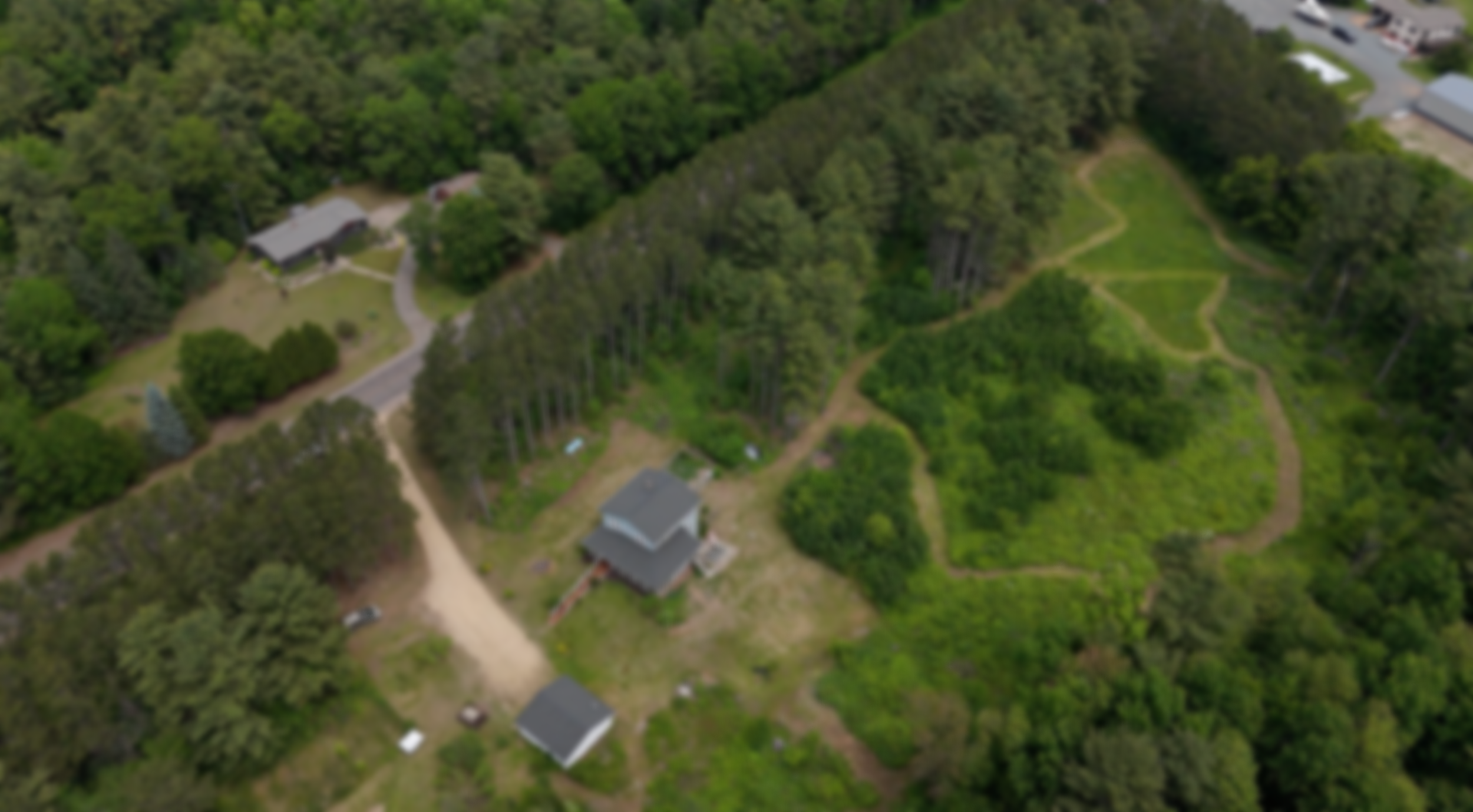 An aerial view of a residential area surrounded by dense green trees, with a house, a smaller building, and several pathways visible.