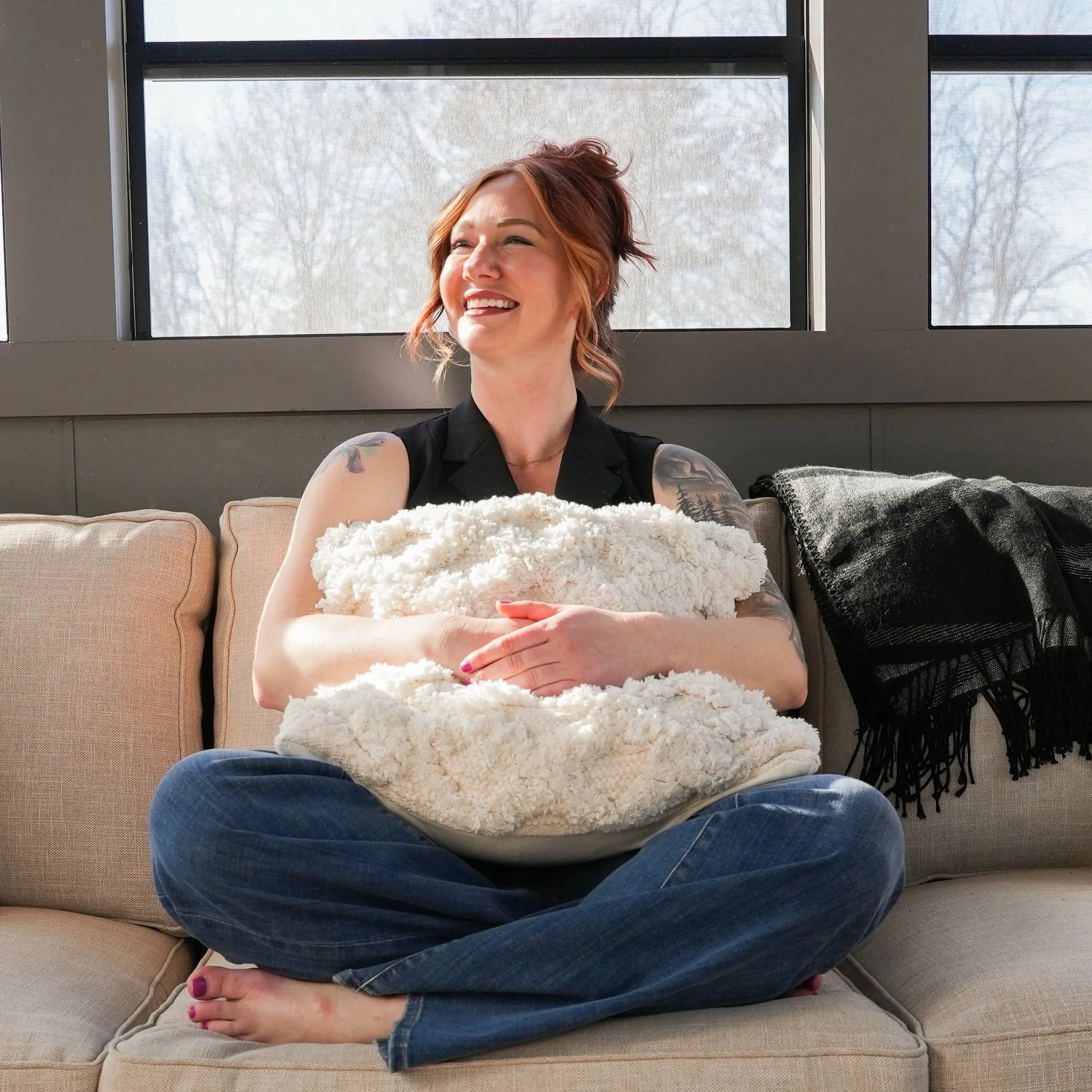 A woman with red hair and tattoos is sitting cross-legged on a beige couch, holding a fluffy pillow and smiling. She is wearing a black sleeveless top and blue jeans. Behind her, large windows show a snowy, tree-covered landscape, and there is a black and gray blanket on the right side of the couch.