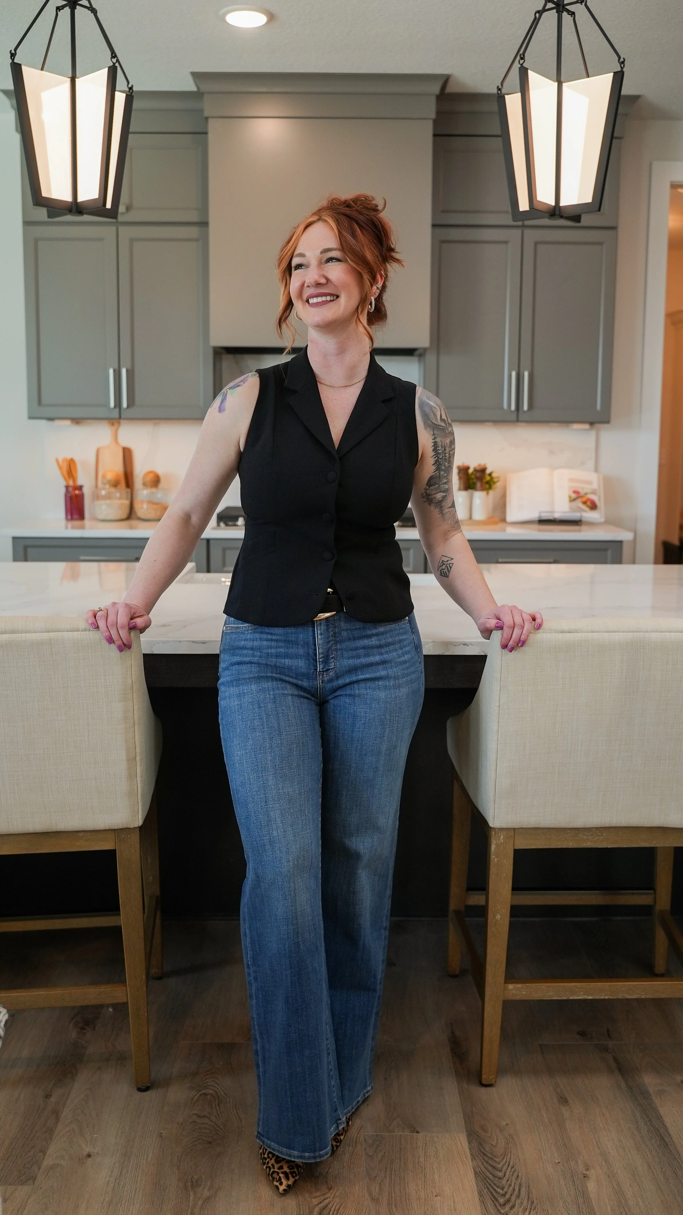 A woman with red hair and tattoos, wearing a black sleeveless top and blue jeans, standing in a modern kitchen with beige chairs and a marble island.