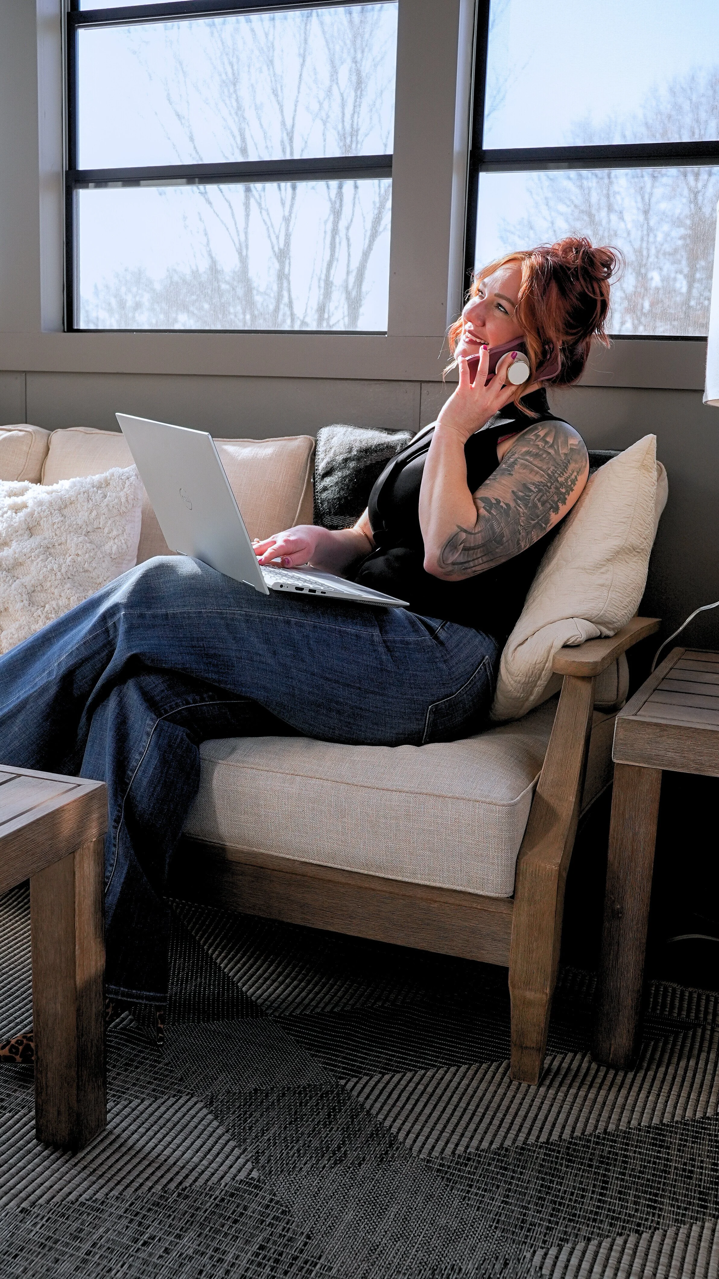 A woman with red hair, tattoos on her arm, wearing a black sleeveless top and jeans, sitting on a beige couch in front of a large window, talking on the phone while using a silver laptop.