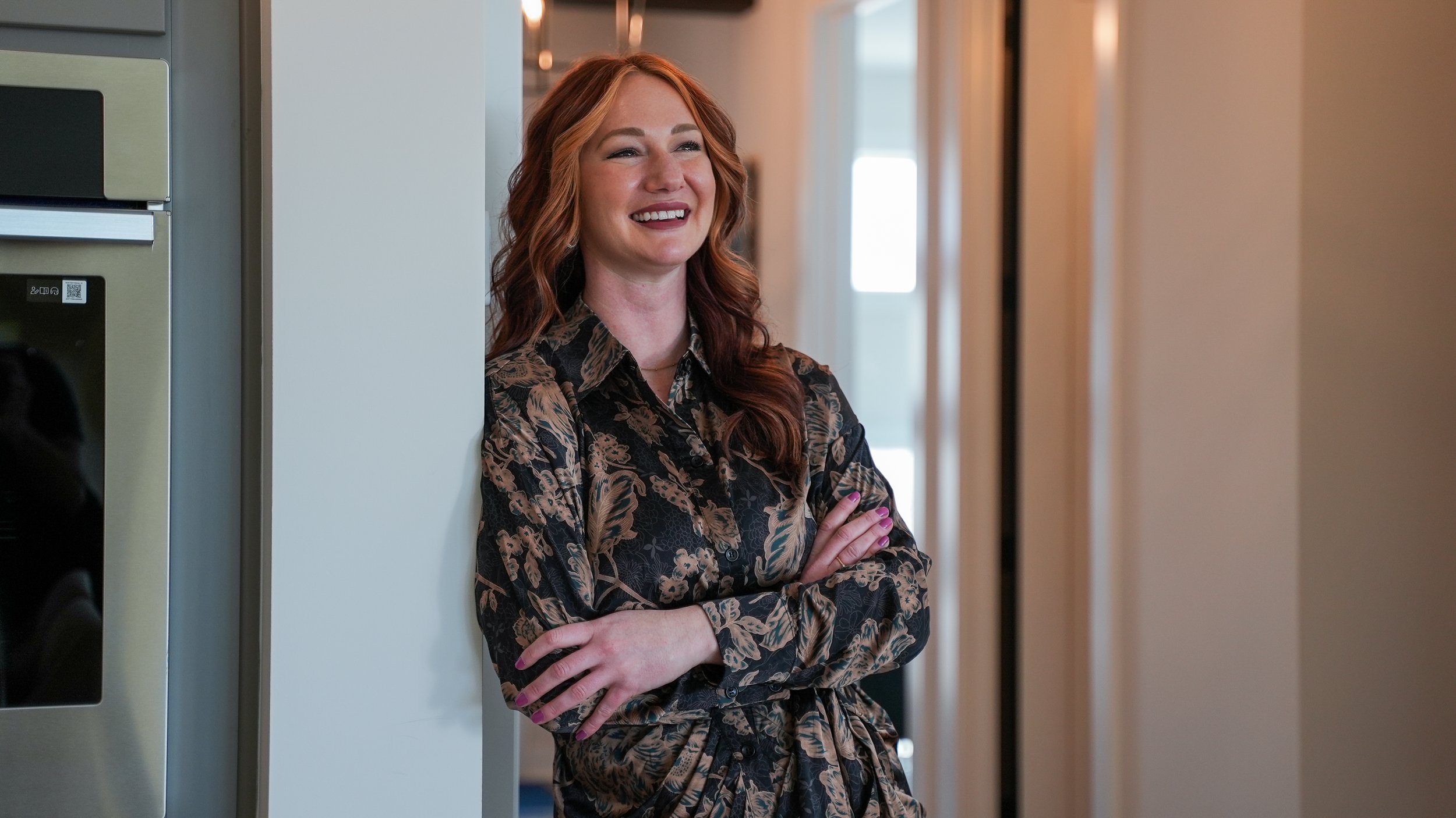 A woman with red hair smiling and crossing her arms, standing indoors near a vending machine and a hallway.