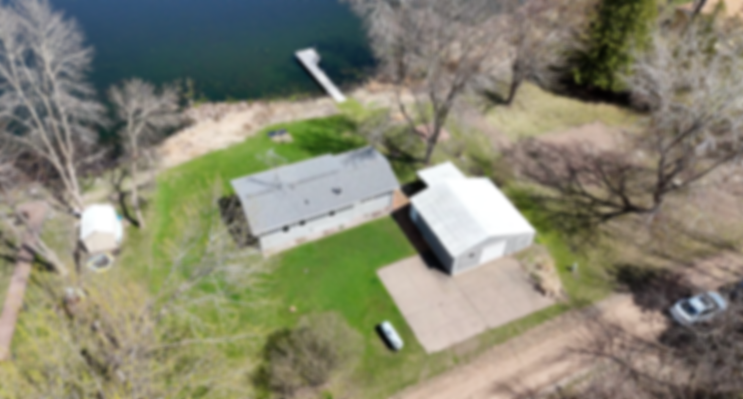 Aerial view of a house with a shed, a lawn, trees, a covered boat dock, water, and parking area in a rural setting.