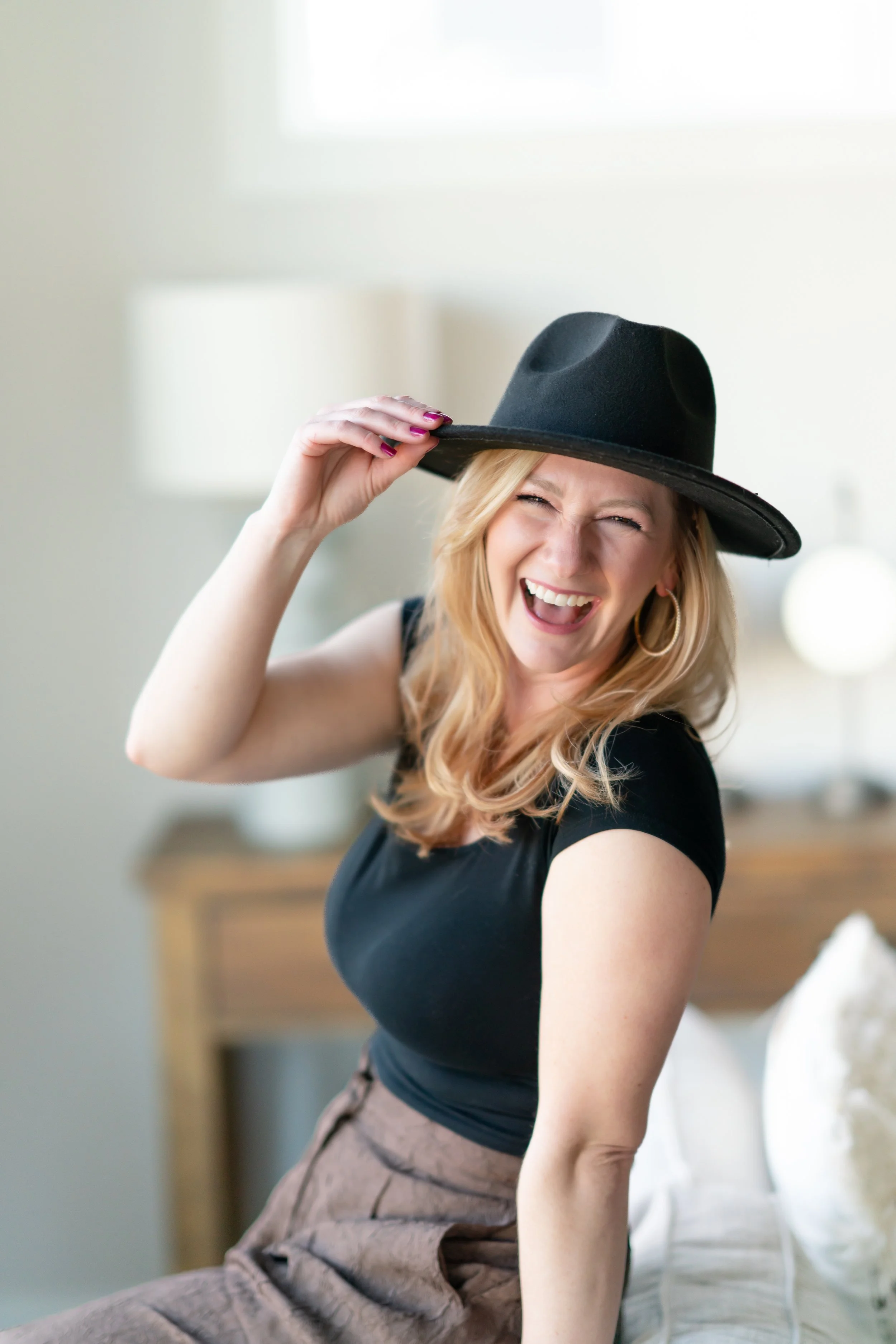 A woman with long blonde hair, wearing a black hat, black shirt, and brown pants, smiling and laughing while holding her hat with one hand in a bright room.