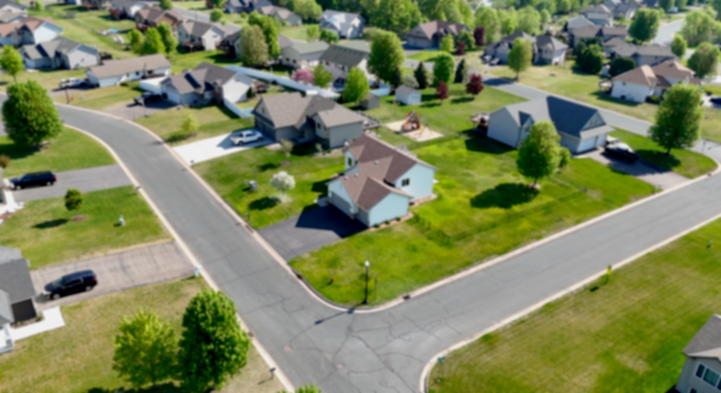 Aerial view of a suburban neighborhood with houses, green lawns, trees, paved roads, and parked cars.