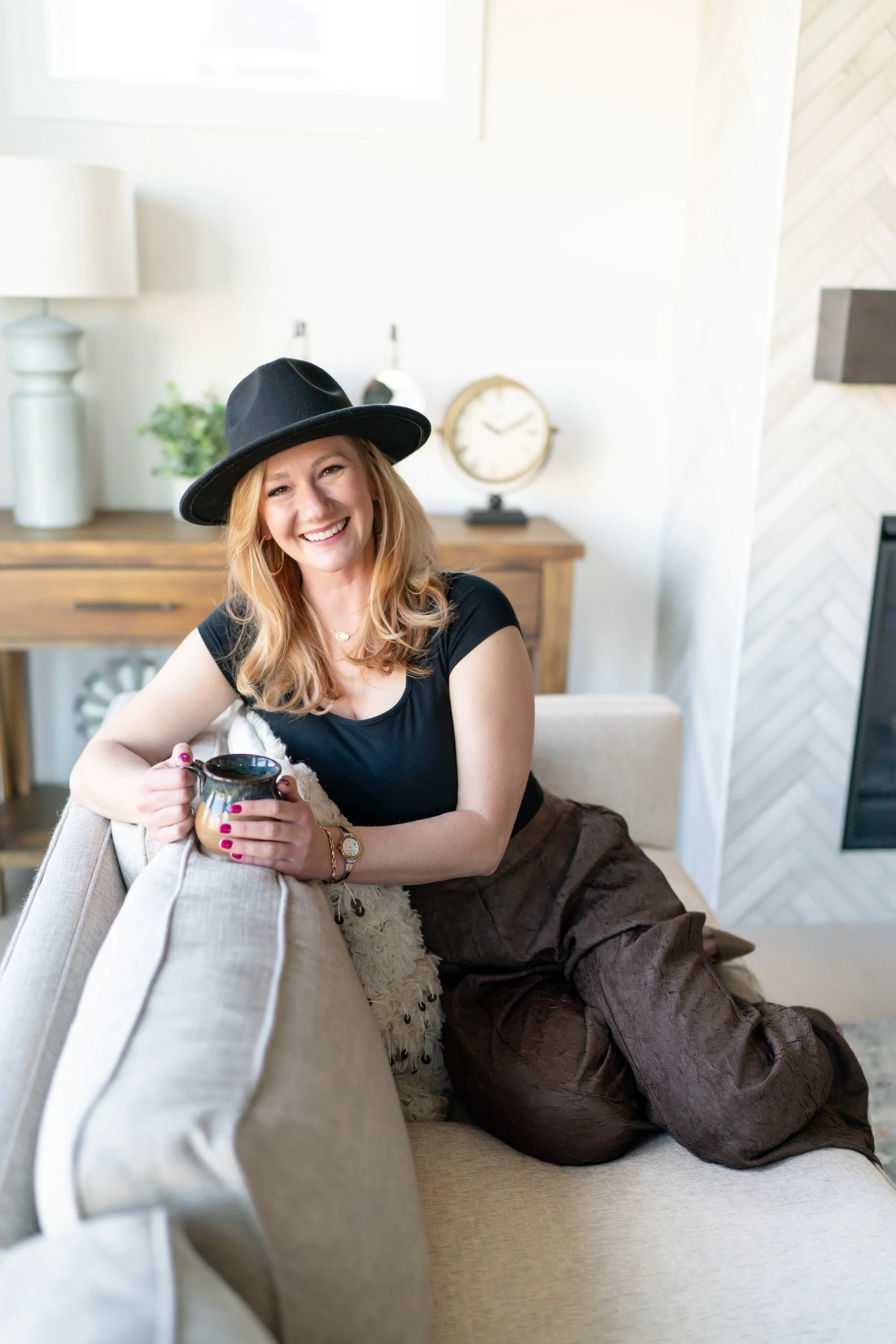 A woman wearing a black hat, black top, and brown pants, sitting on a beige sofa in a bright living room, smiling and holding a mug.
