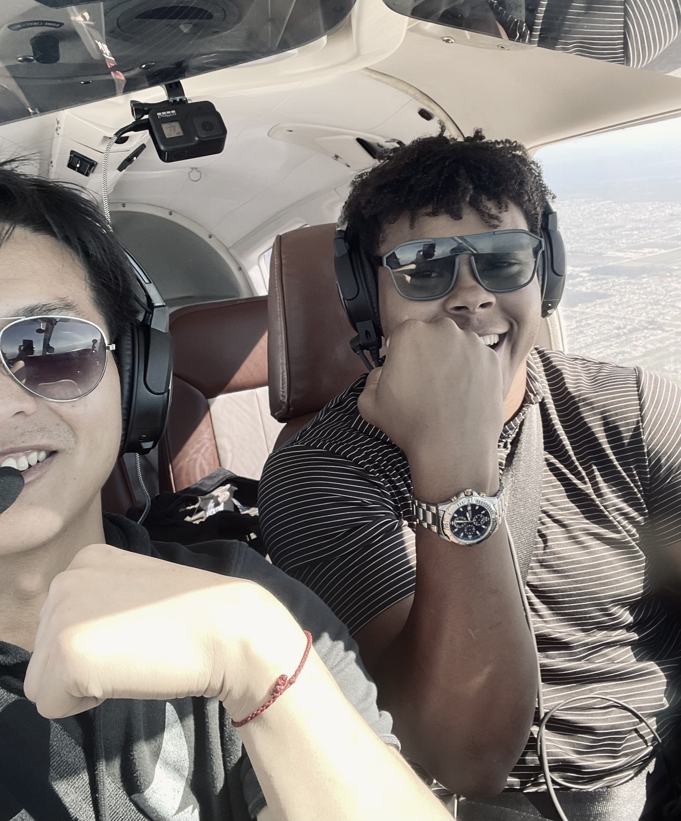 Two men wearing headphones and sunglasses inside an airplane cockpit, one smiling and the other flexing an arm with a watch, with a scenic view outside the window.
