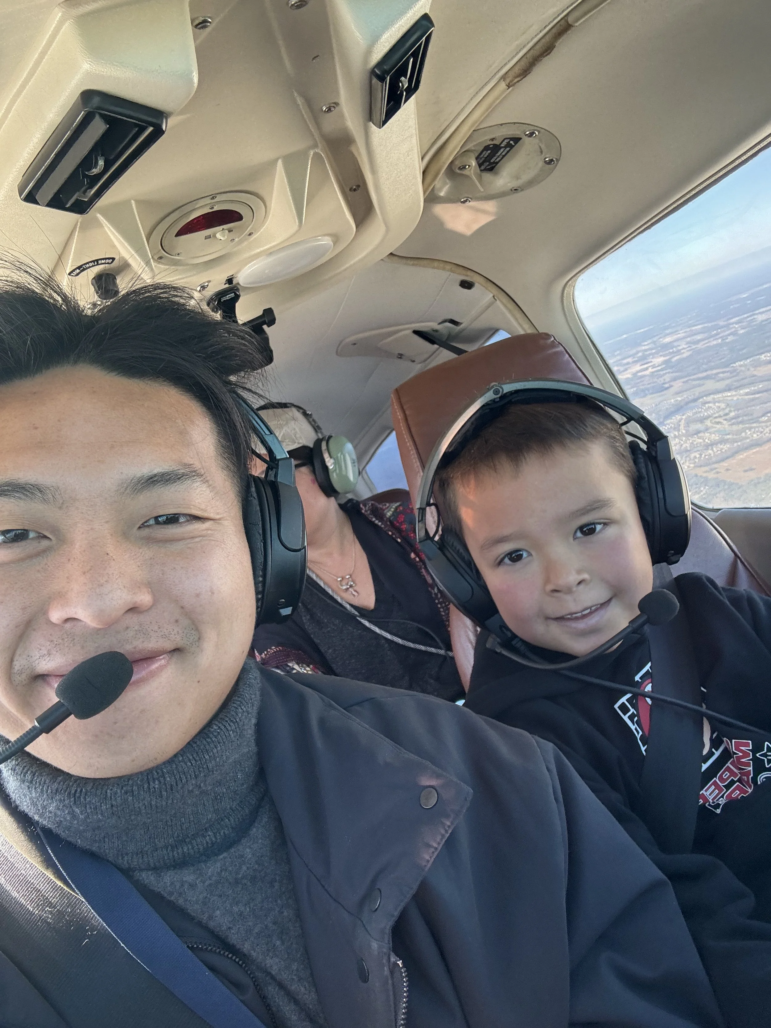 A man and a young boy flying in a small airplane, both wearing headsets, with a woman in the background. The inside of the airplane cockpit is visible with controls and a window showing the sky and landscape outside.