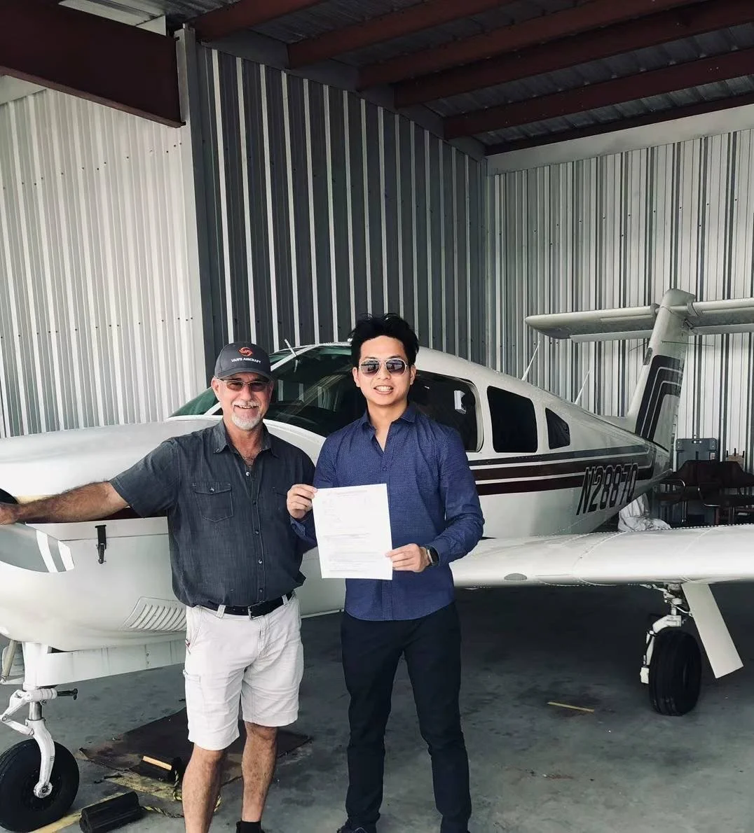 Two men standing in front of a small airplane inside a hangar. One man is holding a document and smiling, both wearing sunglasses. The airplane is white with black and gray stripes, and the hangar has metal walls and a ceiling with metal beams.
