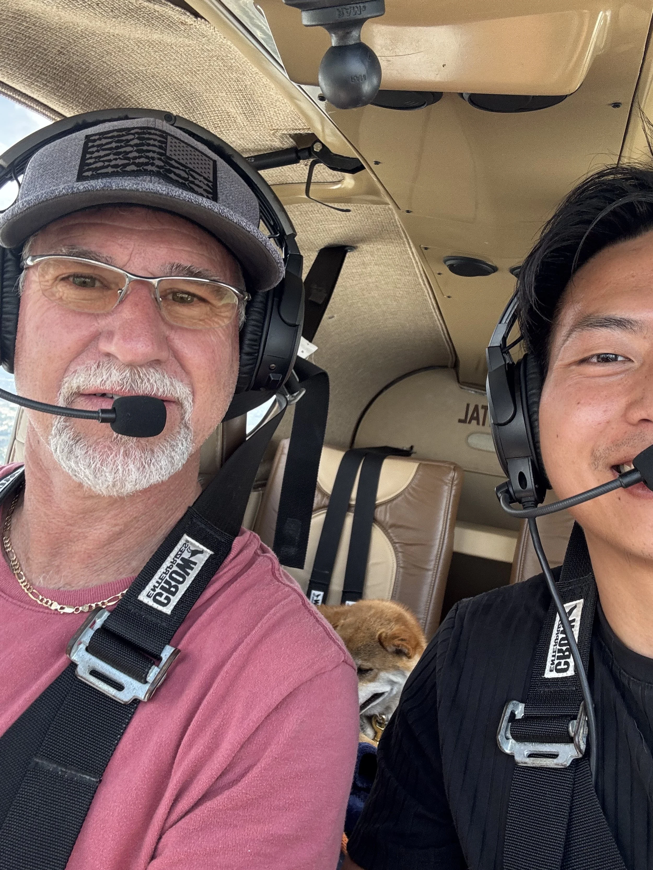 Two men in an airplane cockpit wearing headsets, with a dog sleeping in the backseat.