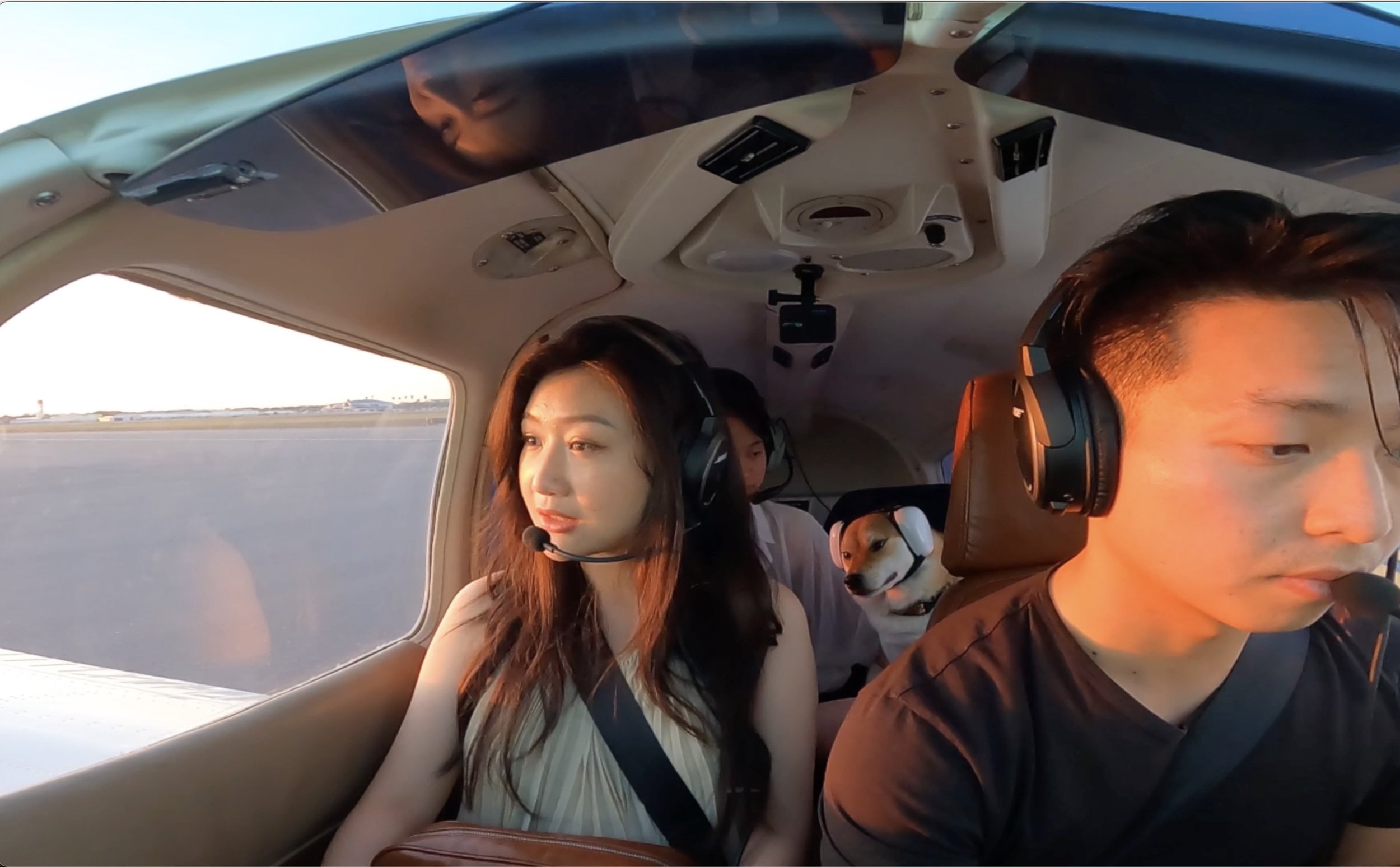 Four people and a dog inside a small aircraft cockpit, with an outdoor runway visible through the side window, during sunset.