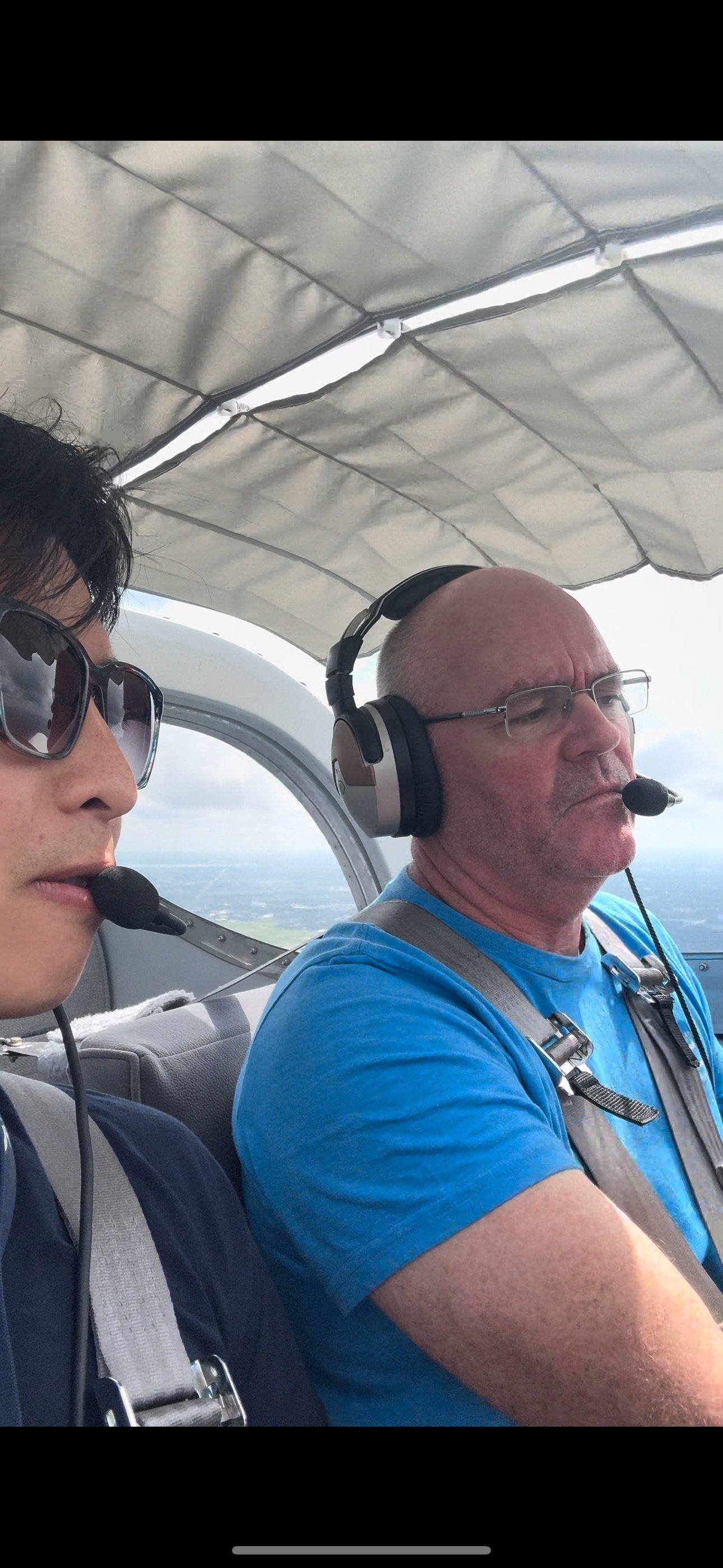 Two men flying in a small plane, both wearing headsets with microphones. The man on the left is wearing sunglasses, and the man on the right is wearing glasses and a blue shirt.