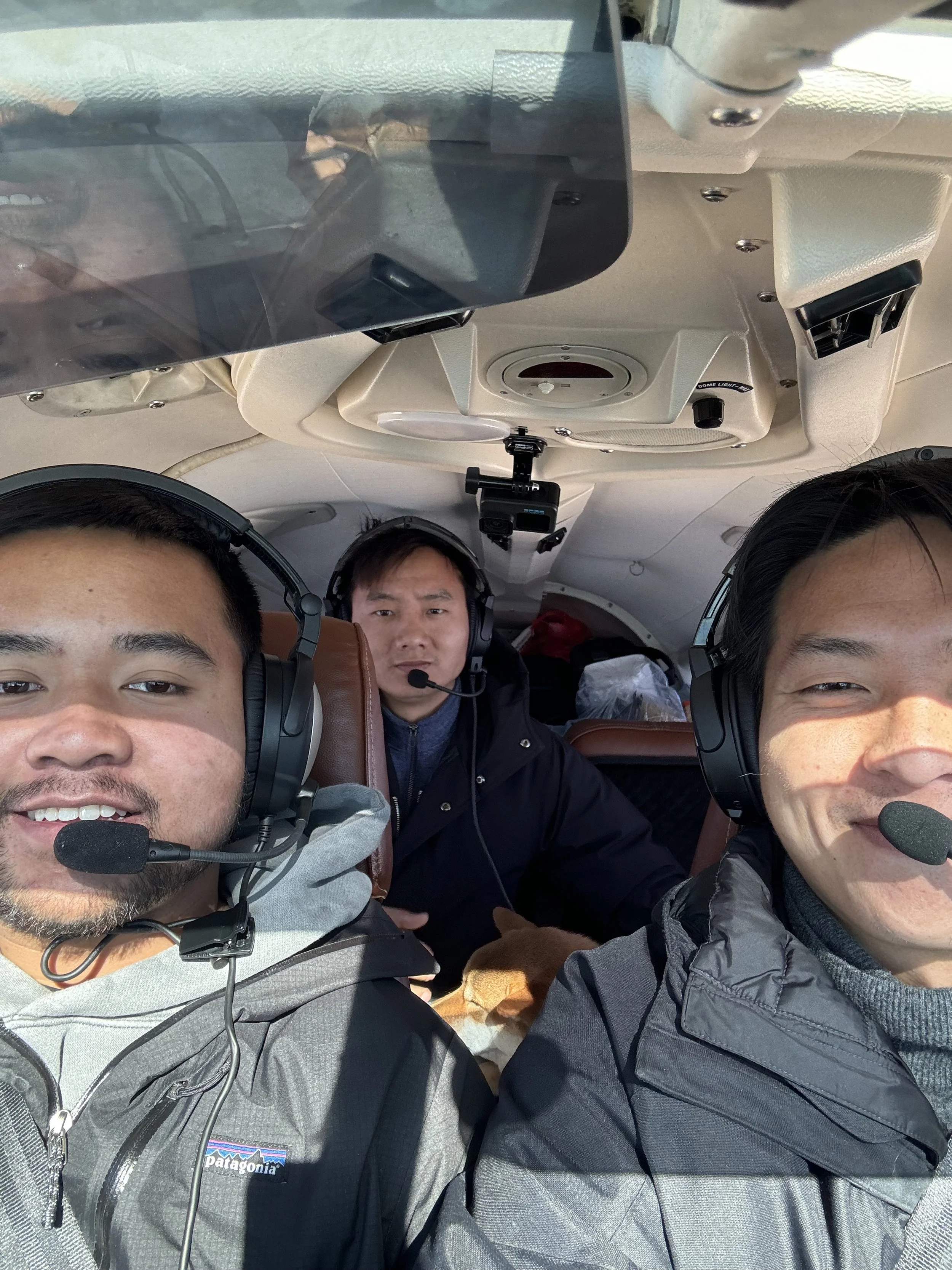 Three men wearing headsets with microphones inside an airplane cockpit, smiling for a selfie.