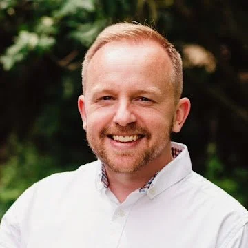Headshot of a smiling man with short hair and a beard, wearing a white collared shirt, outdoors with greenery in the background.