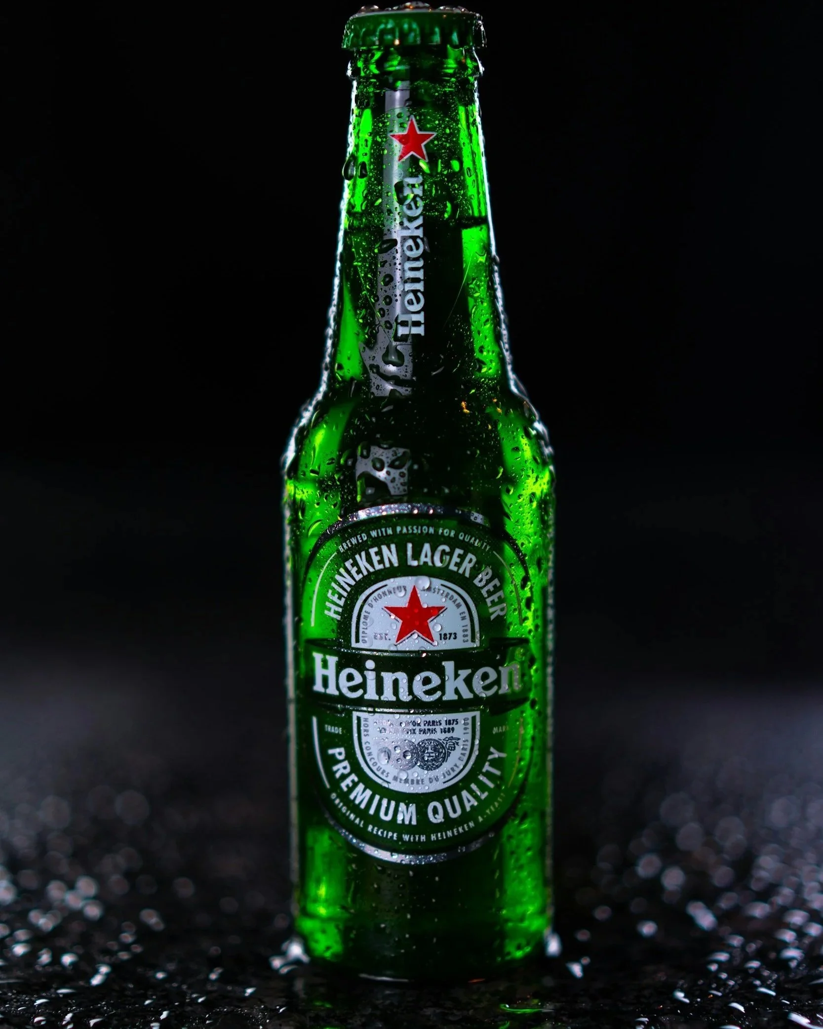 A green bottle of Heineken beer covered in water droplets, standing on a wet surface against a black background.
