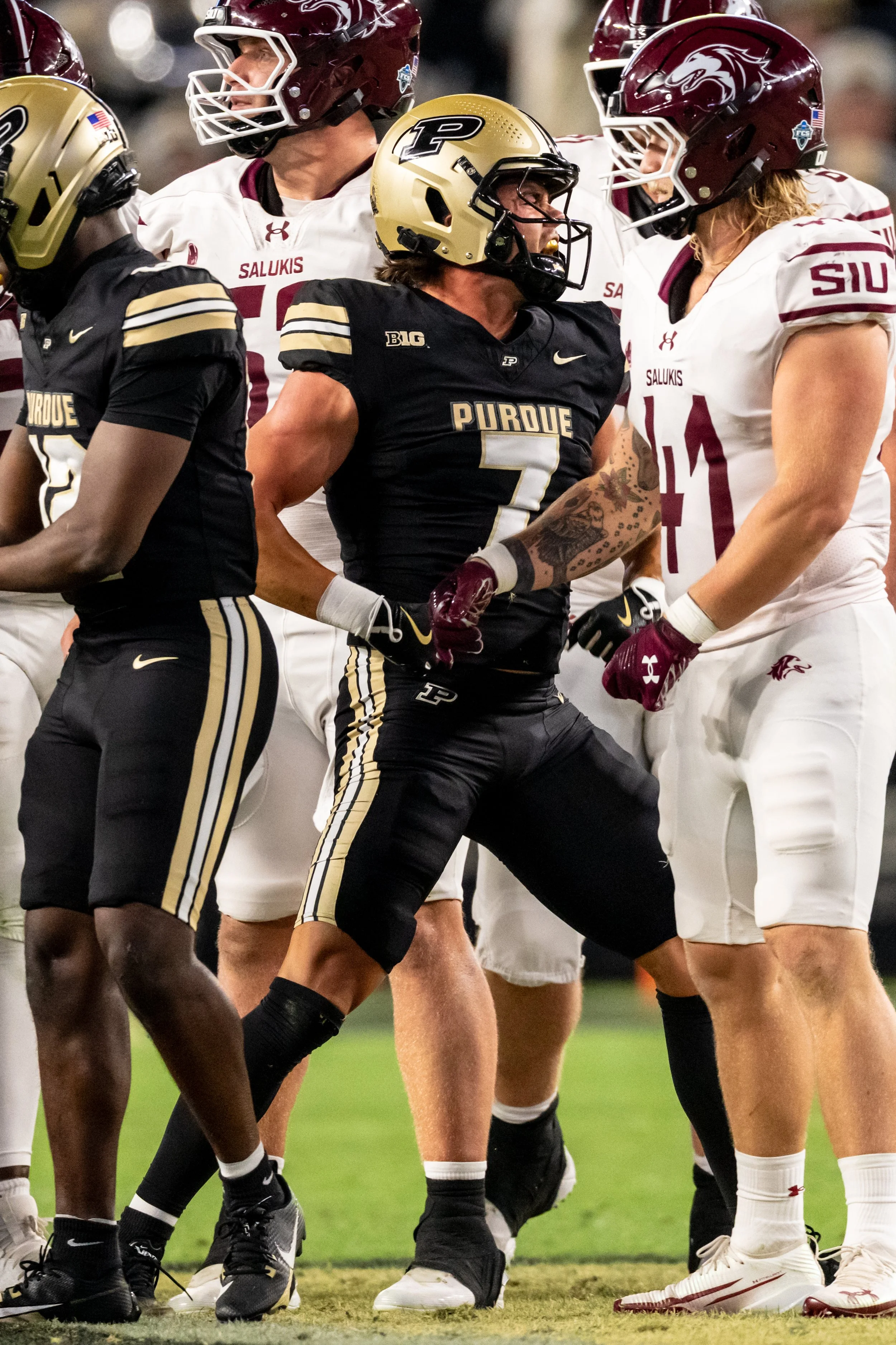 American football players from Purdue and Indiana State in a game, wearing helmets and uniforms, engaging in a competitive moment on the field.