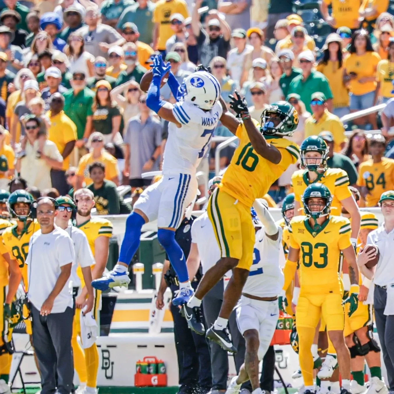 An American football game with a player from the team in white and blue leaping to catch the ball, while a player from the team in yellow and green attempts to block him. The background shows a large crowd of spectators, many wearing yellow, green, and blue colors.