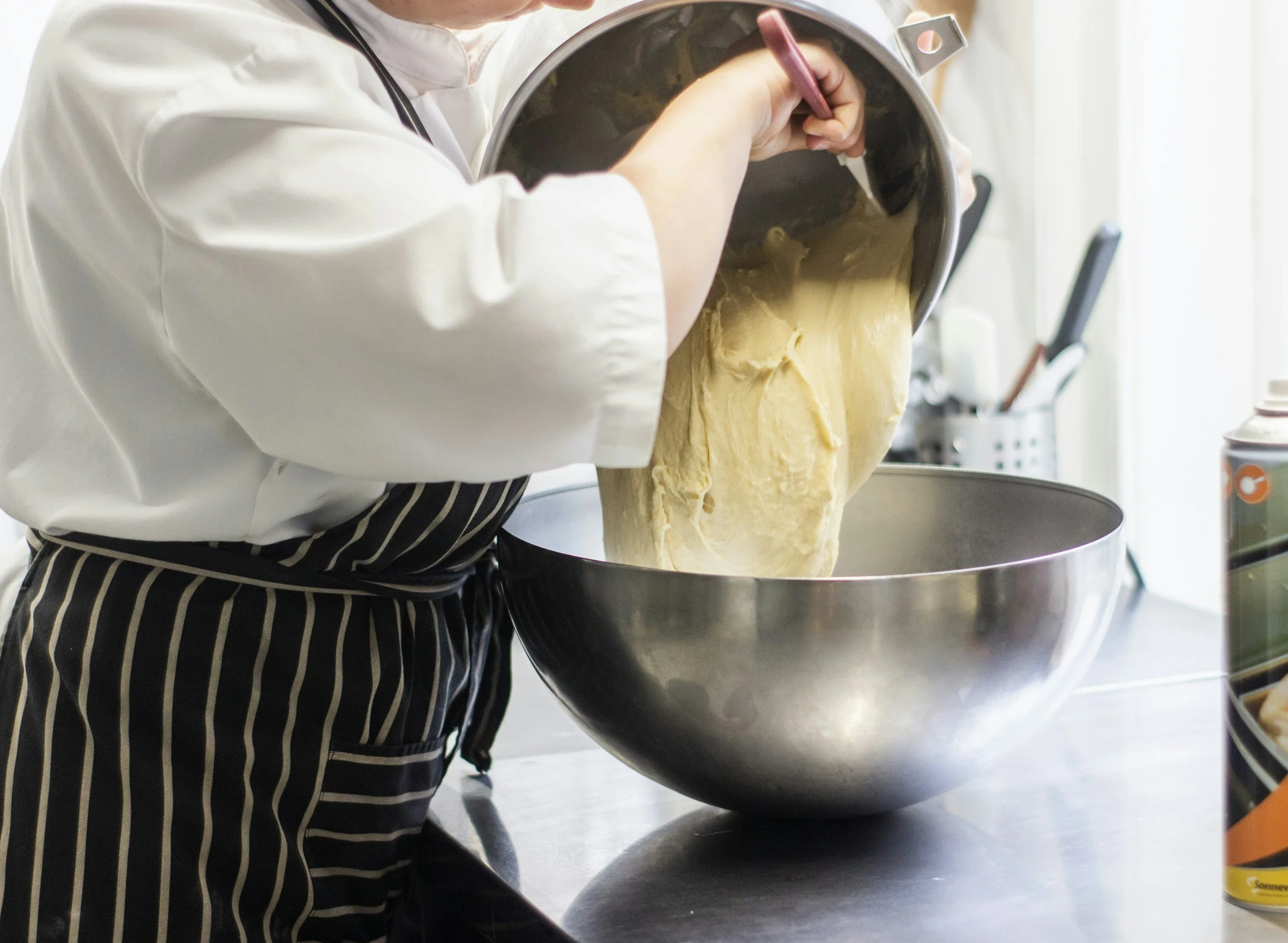 Person pouring dough from a mixing bowl into a large metal bowl in a kitchen.